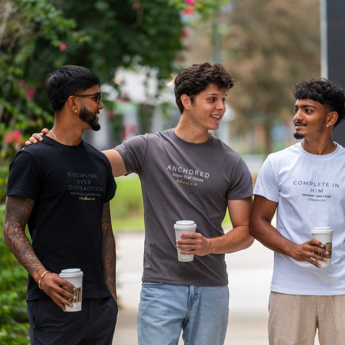 Three young men standing outdoors on a sidewalk, each wearing a graphic t-shirt. The man on the left wears a black t-shirt with gold text, the man in the middle wears a gray t-shirt with white text, and the man on the right wears a white t-shirt with black text.