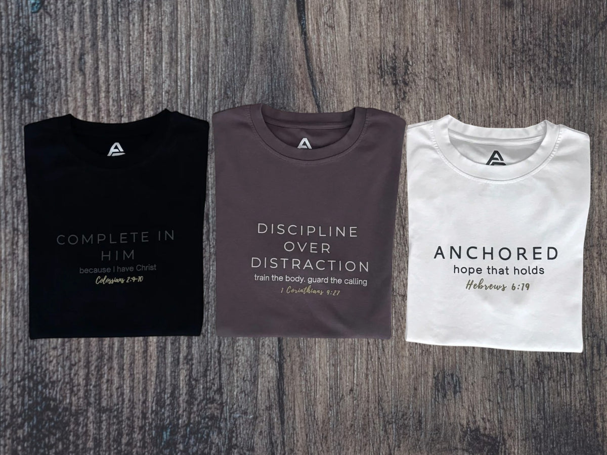 Three folded graphic t-shirts in black, dark gray, and white, laid out side-by-side on a dark wood background. Each shirt features a minimalist geometric logo on the chest and a scripture quote printed below.