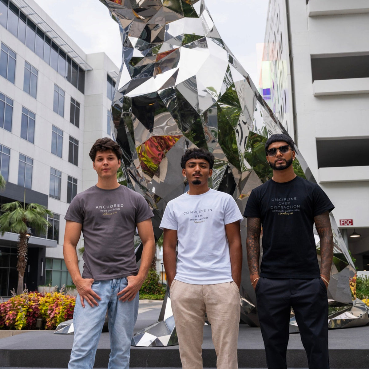 Three men modeling ADM Driven shirts in charcoal Anchored, white Complete In Him, and black Discipline Over Distraction, standing in front of a modern reflective sculpture.