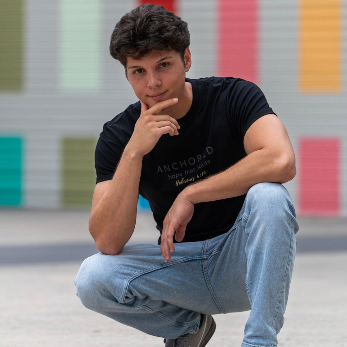 A young man crouching in front of a colorful striped wall, wearing a black graphic t-shirt with 'ANCHOR5' printed on the chest and light blue jeans.