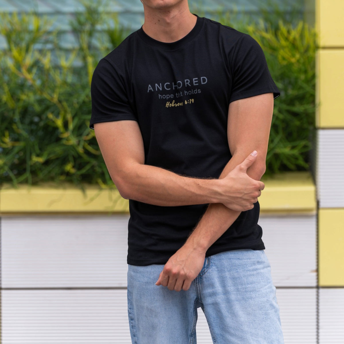 Man wearing the black Anchored men's shirt by ADM Driven, featuring the message inspired by Hebrews 6:19, paired with a modern outdoor setting.