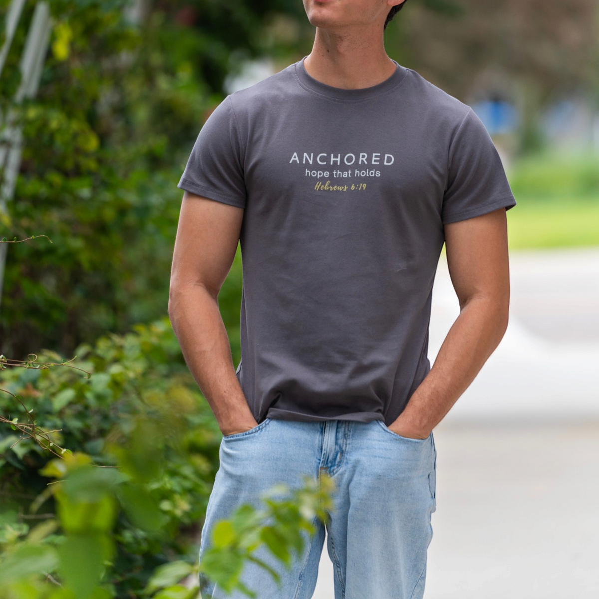 A man wearing a dark gray crewneck t-shirt with the text 'ANCHORED hope that holds Philippians 4:19' printed on the chest, paired with light blue jeans, standing outdoors with blurred green foliage in the background.