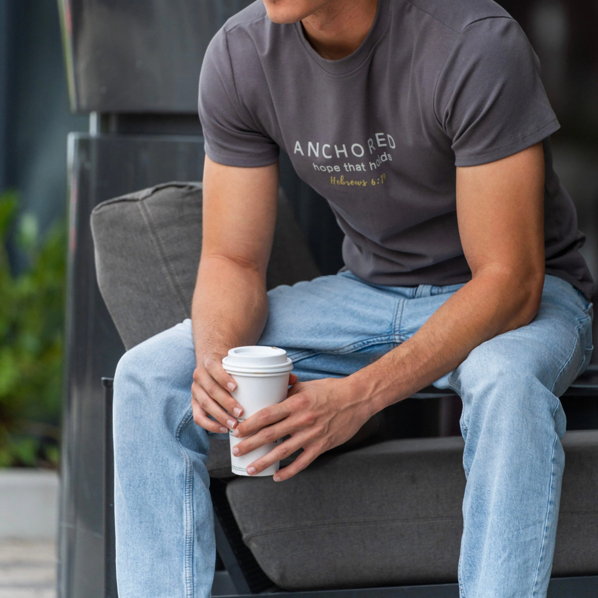 A man sitting on a dark gray outdoor bench wearing a dark gray t-shirt with the text 'ANCHORED hope that matters' in white and yellow, paired with light blue jeans, holding a white paper coffee cup.