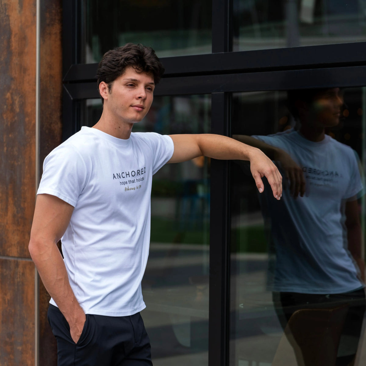 Man wearing the white Anchored men's shirt by ADM Driven, leaning against a modern glass building, with the chest design inspired by Hebrews 6:19.