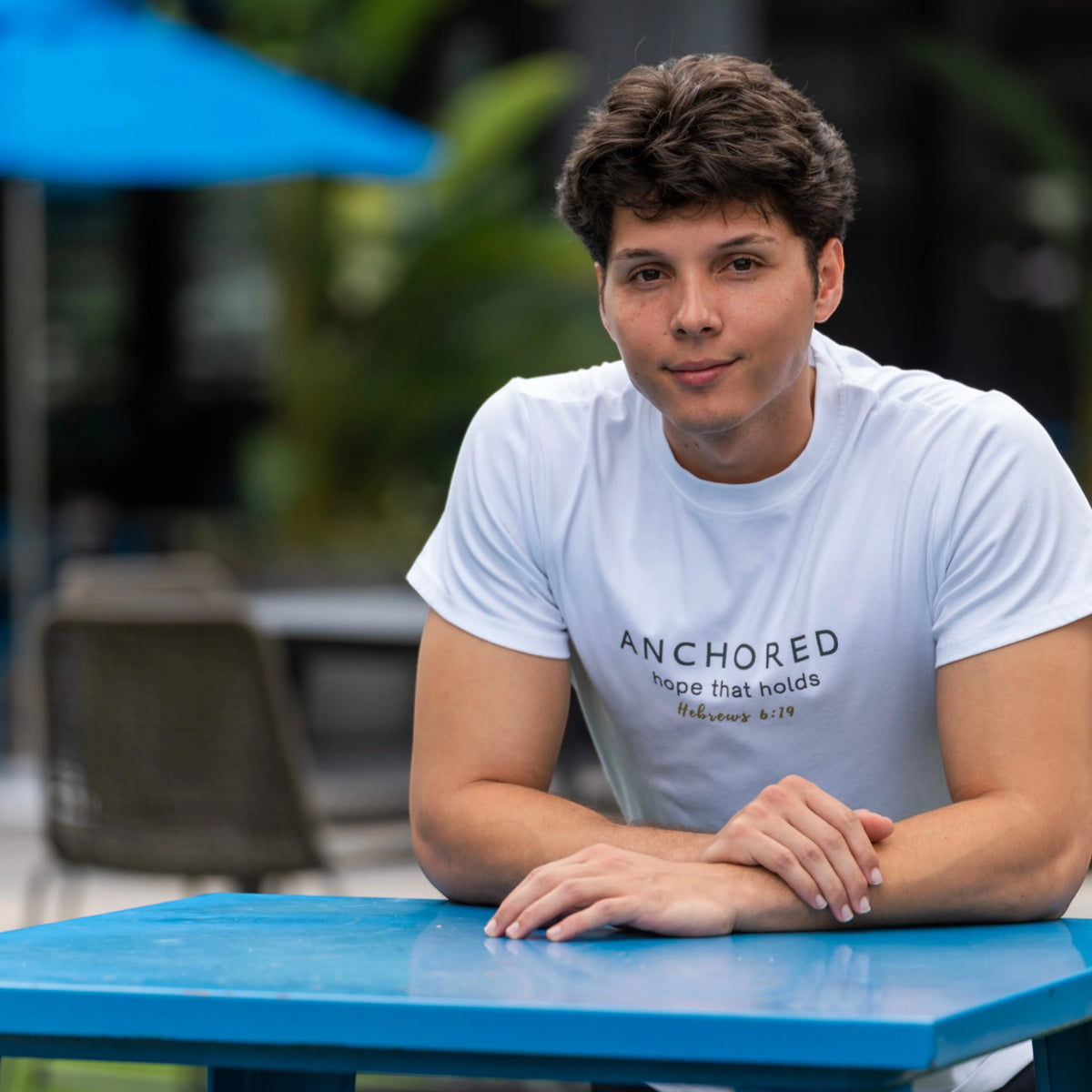 A man wearing a white graphic t-shirt with the text 'ANCHORED hope that holds Matthew 6:19' sitting at a bright blue outdoor table.