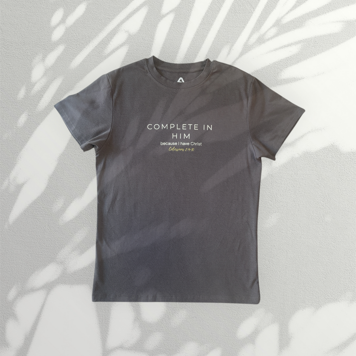 Charcoal grey Complete In Him men's shirt by ADM Driven featuring a white and gold chest design, laid flat against a light background with soft leaf shadows.