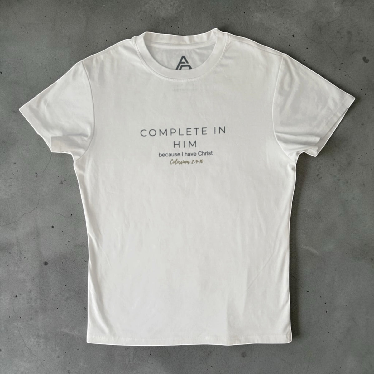 A white short-sleeve crewneck t-shirt with black printed text on the chest reading 'COMPLETE IN HIM because I Have Christ' and 'Colossians 2:10', laid flat on a textured gray concrete surface.