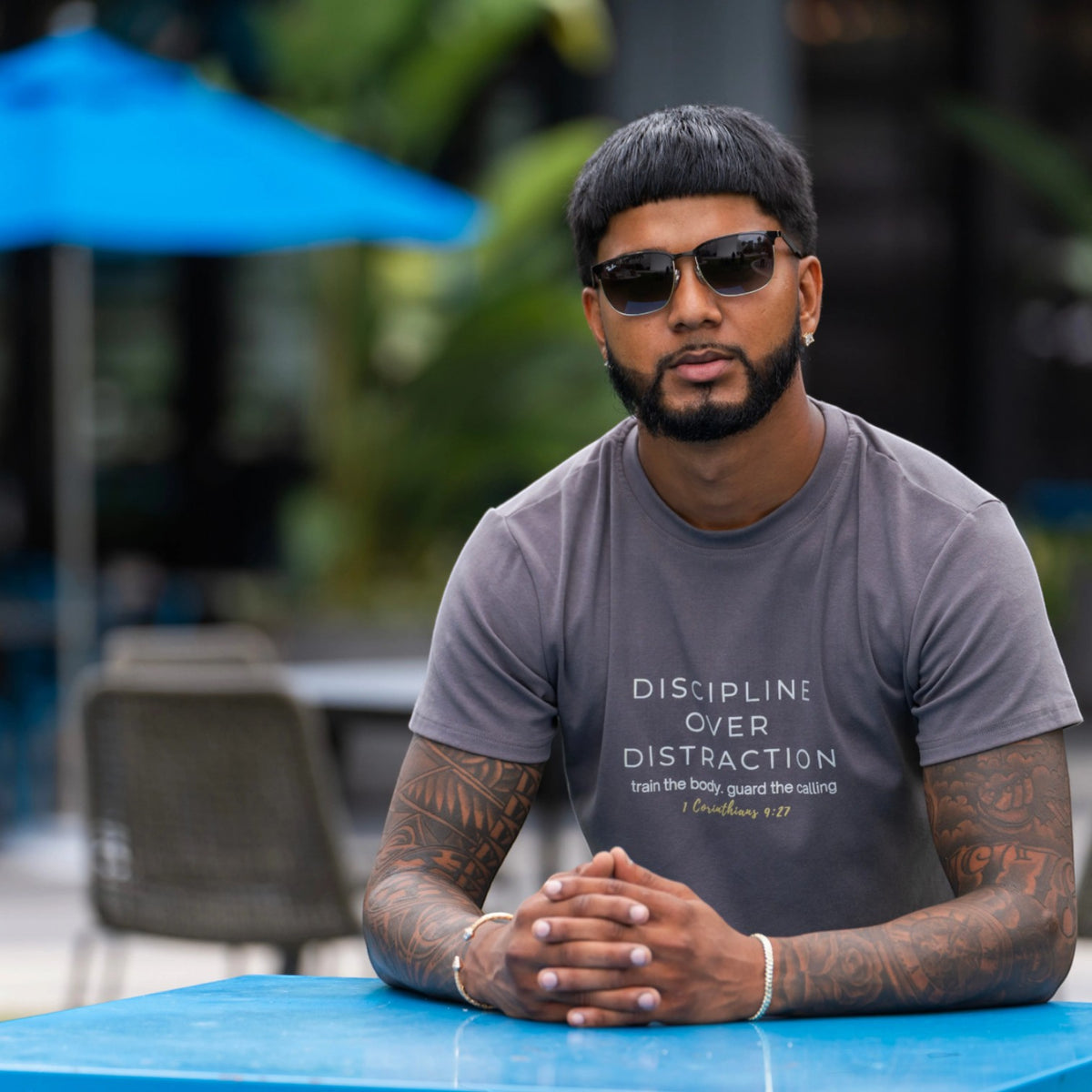 A man with tattoos and sunglasses sits at a blue table outdoors, wearing a gray t-shirt with the text 'DISCIPLINE OVER DISTRACTION' printed on the front.