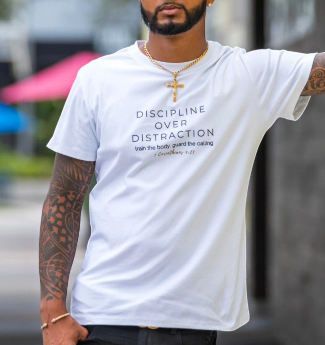 A man wearing a white graphic t-shirt with the text 'DISCIPLINE OVER DISTRACTION' and 'train the body guard the calling 1 Peter 4:12' printed in navy blue. The photo is taken from the chest up, showing tattooed arms and a gold cross necklace, with a blurred outdoor background.