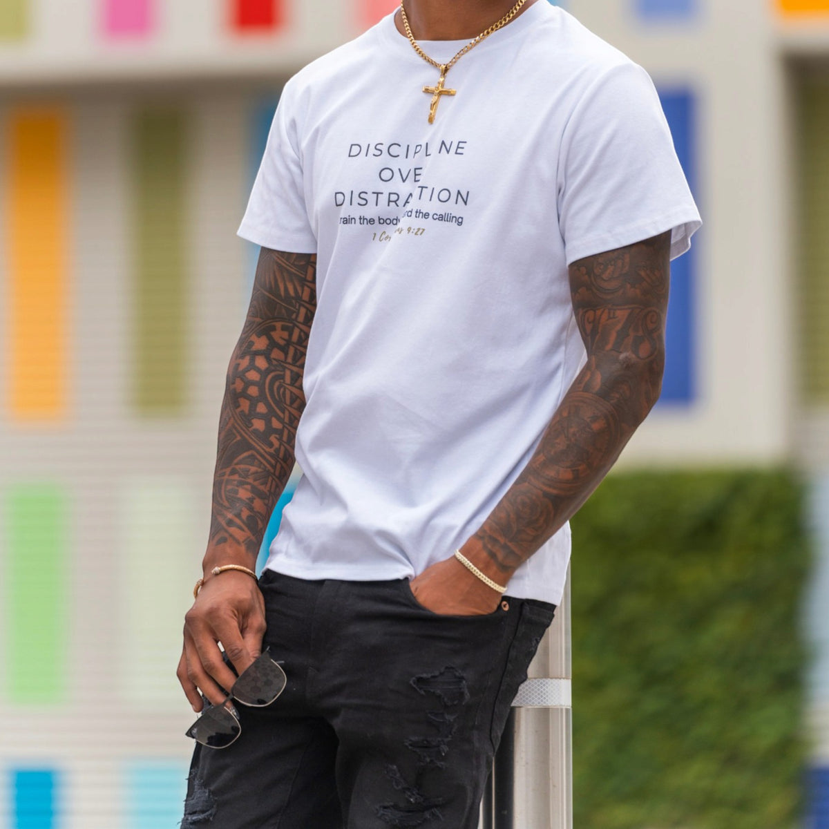 A man wearing a white graphic t-shirt with the text 'DISCIPLINE OVE DISTRACTION' and 'use the step * the stepping' printed in black and gold. He is also wearing black distressed jeans, a gold chain necklace with a cross, and holding sunglasses.